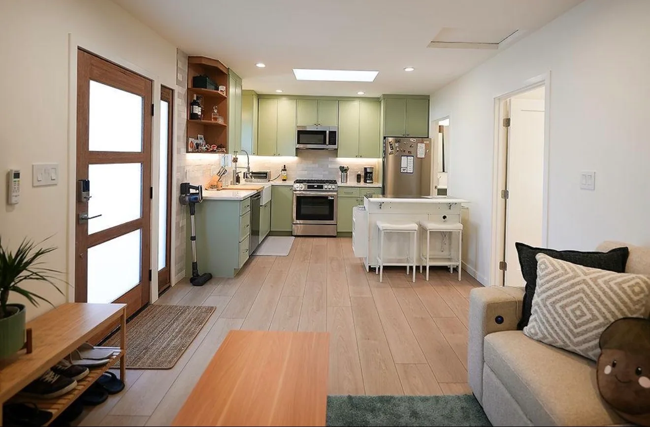 Interior of a small open-plan dwelling with a galley kitchen featuring green cabinets, stainless steel appliances, a wood-floor living area with a sofa, and a glass-paneled entry door.