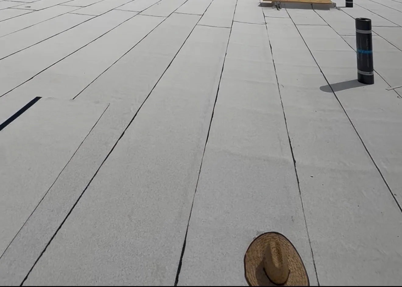 A flat commercial roof surface covered with light-colored membrane seams, with a roll of roofing material and a straw hat resting on the surface.