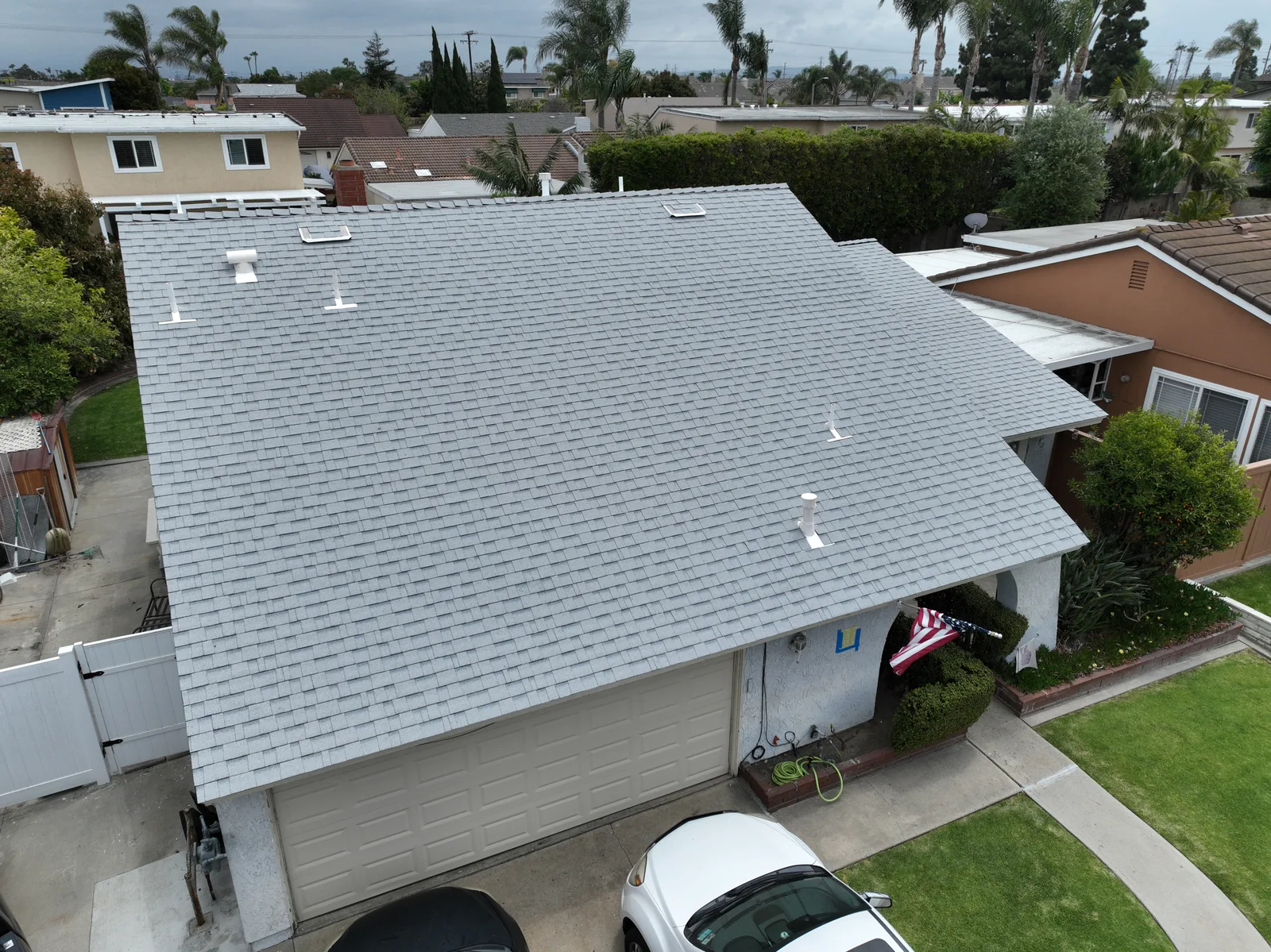 Aerial view of a single-story house with a newly installed light gray shingle roof, featuring vents and a skylight, with a garage and parked cars below.