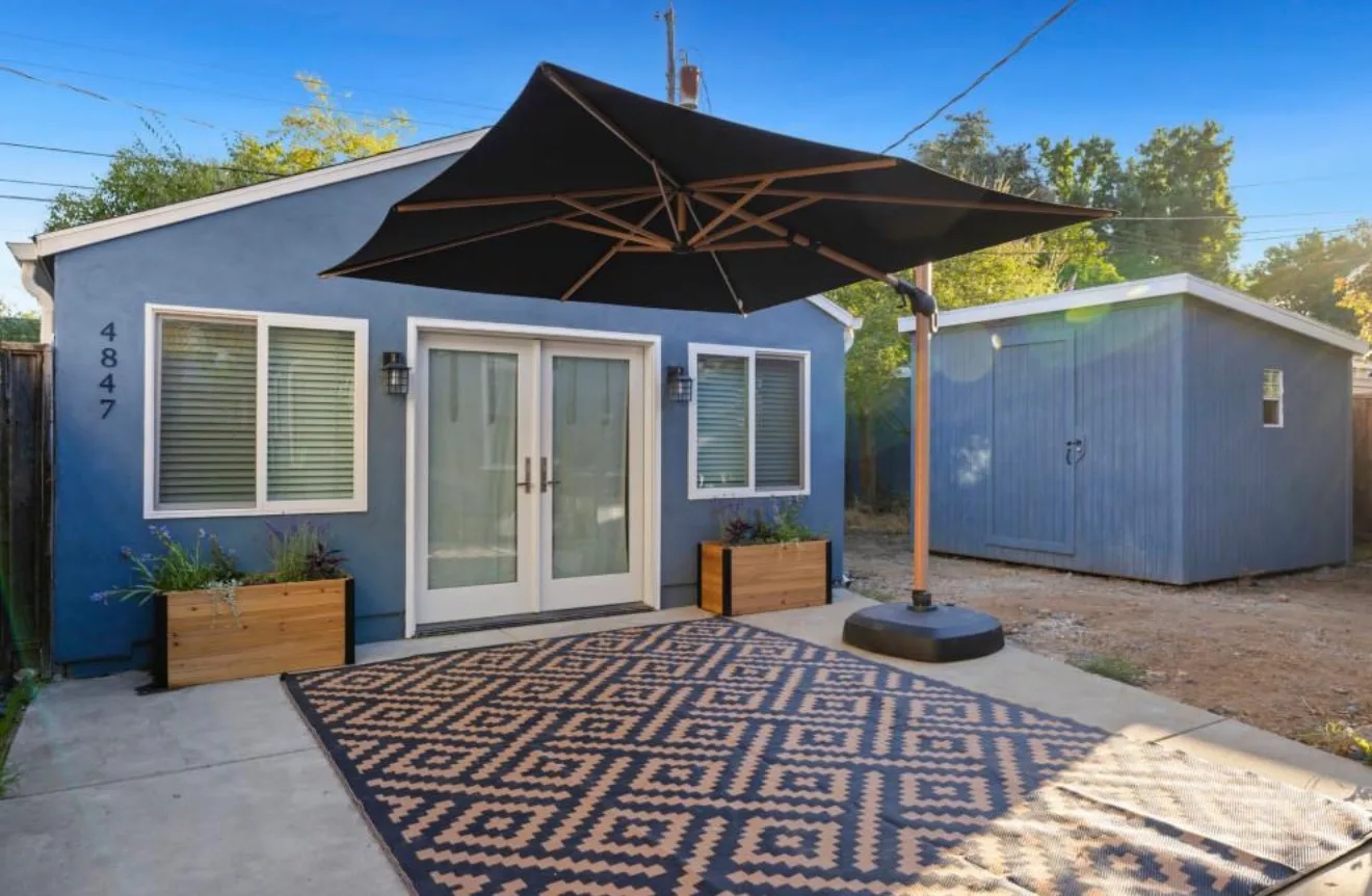 A blue stucco single-story home with white-trimmed French doors and windows, a patterned outdoor rug on a concrete patio under a large black cantilever umbrella, wooden planter boxes, and a matching blue storage shed to the right.