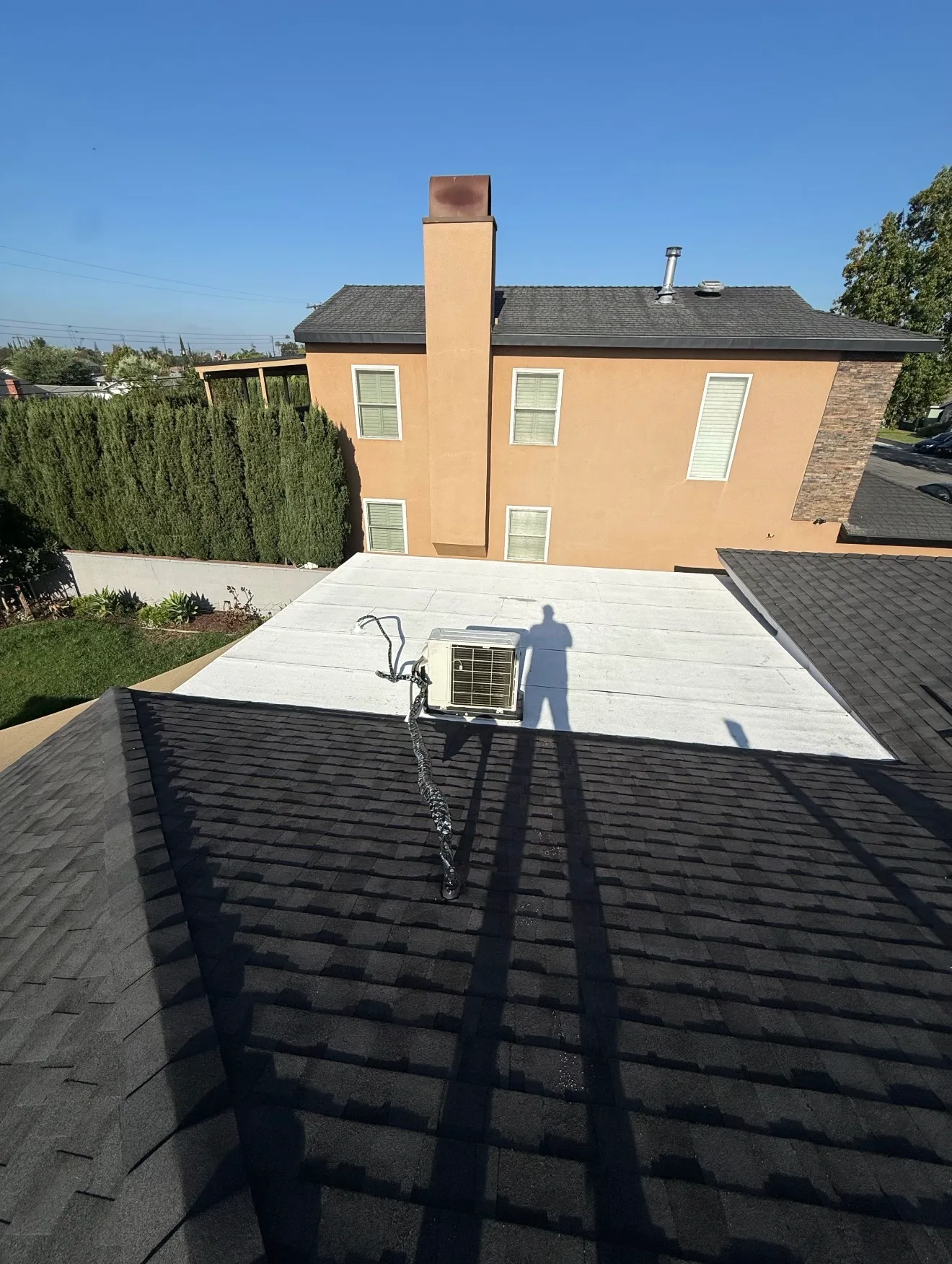 A dark asphalt shingle sloped roof in the foreground next to a flat white low-slope roof holding a mini-split condenser unit, with a two-story stucco house and chimney in the background.